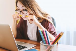 A girl nervously bites her pencil while taking an assessment on her laptop.