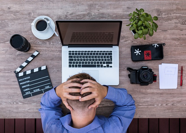 A desk with a laptop, cup, plant, and other creative tools. The man is sitting with his hands on his head.